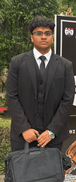 A young man wearing a formal black three-piece suit and glasses, holding a black briefcase.