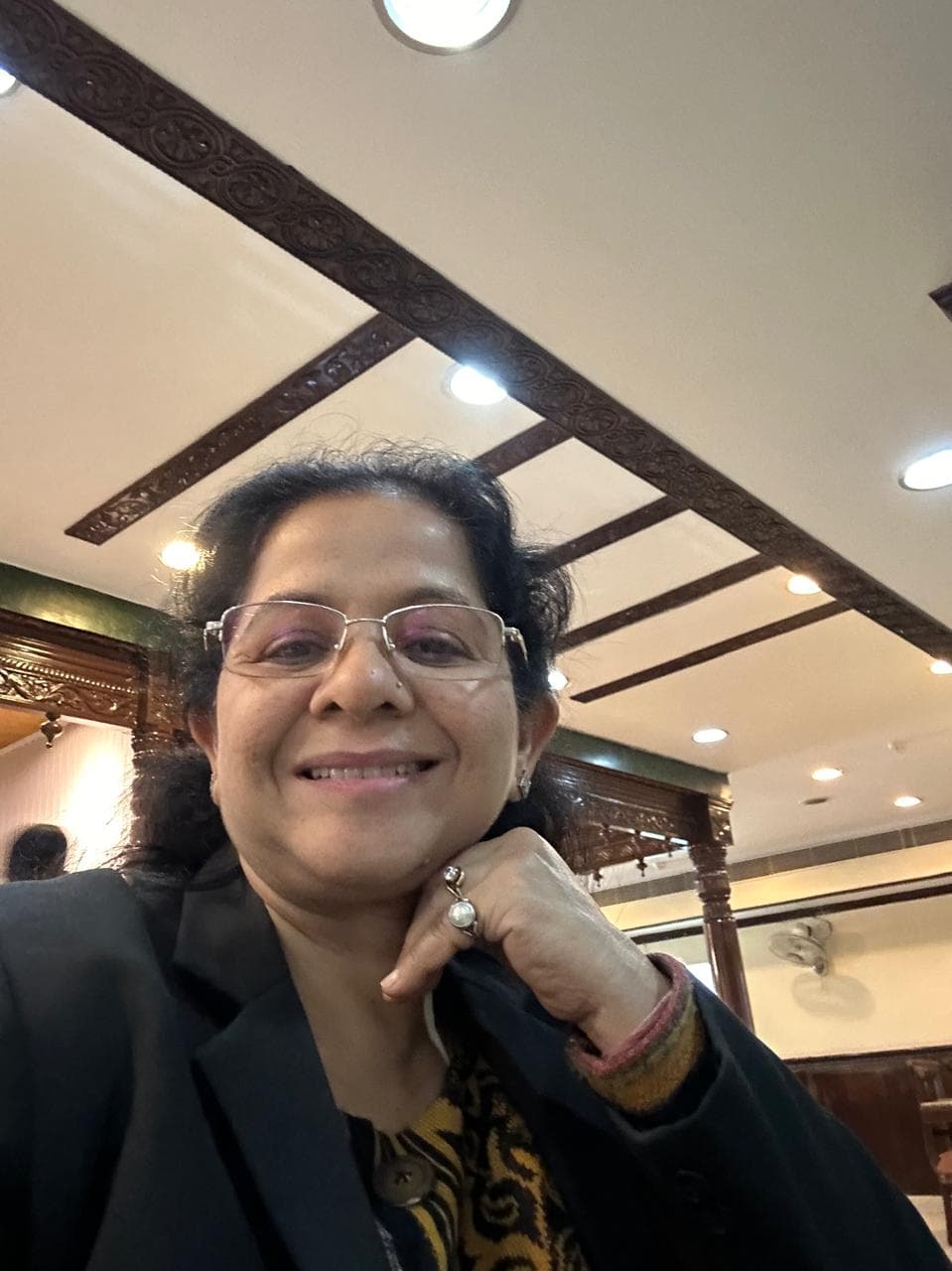 Smiling woman with glasses and a blazer, hand on chin, under a decorative ceiling.